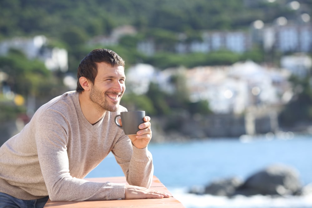 Happy man drinking coffee contemplating views in a balcony on the beach a sunny day